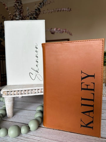 Brown leather cover with 'KALEY' engraved, white cover with 'Shannon' engraved, on a wooden surface.