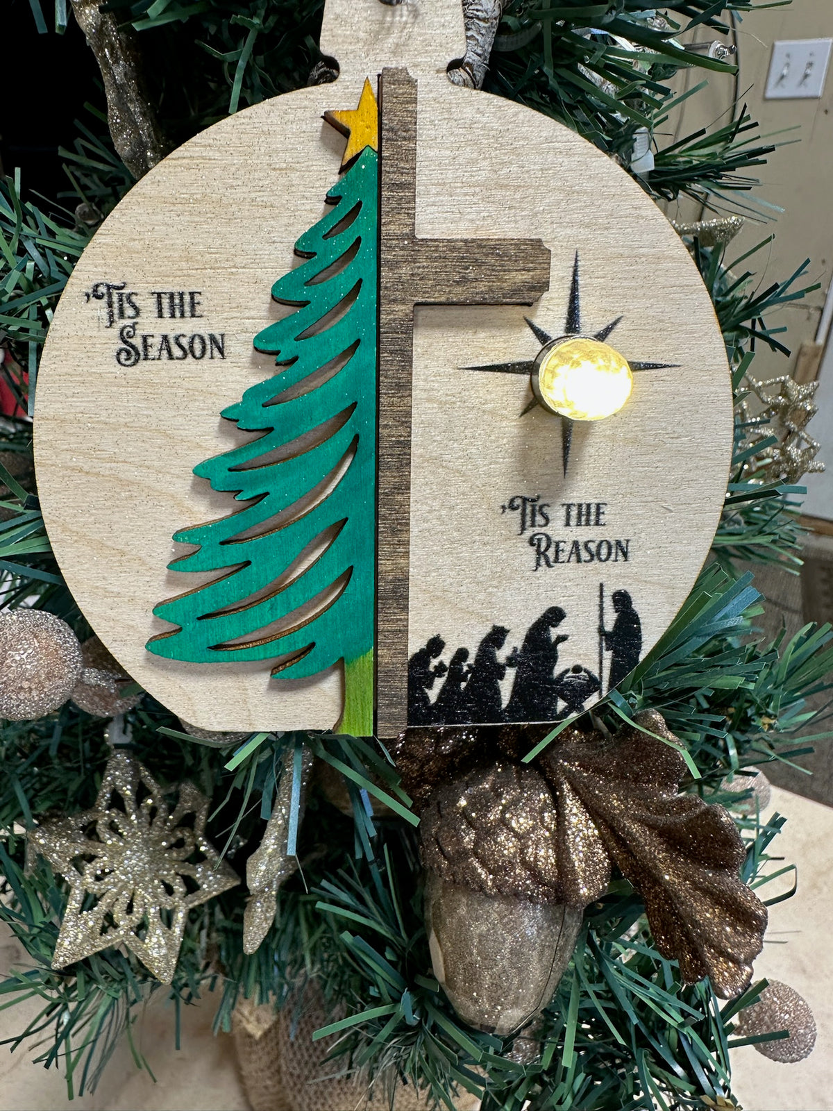 A round wooden Christmas ornament hanging on a green Christmas tree, featuring a split design with a green tree on one side and a cross on the other, plus the phrases 'It's the Season' and 'It's the Reason', a star, and silhouettes of the Holy Family.