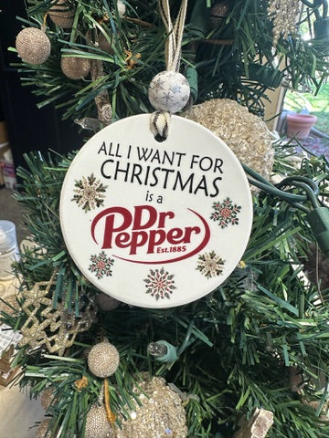 A round white Christmas ornament hanging on a green artificial tree. The ornament has black text reading 'ALL I WANT FOR CHRISTMAS is a' above a red Dr Pepper logo, with small snowflake designs around the border.