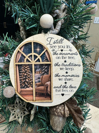 A round wooden Christmas ornament hanging on a green holiday tree. The ornament features a printed illustration of a cozy living room window with a Christmas tree and armchair, alongside the sentimental text: 'I still see you in.