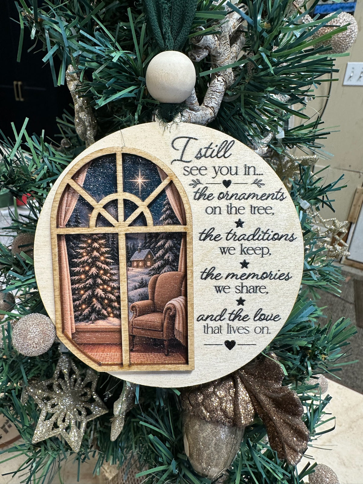 A round wooden Christmas ornament hanging on a green holiday tree. The ornament features a printed illustration of a cozy living room window with a Christmas tree and armchair, alongside the sentimental text: 'I still see you in.