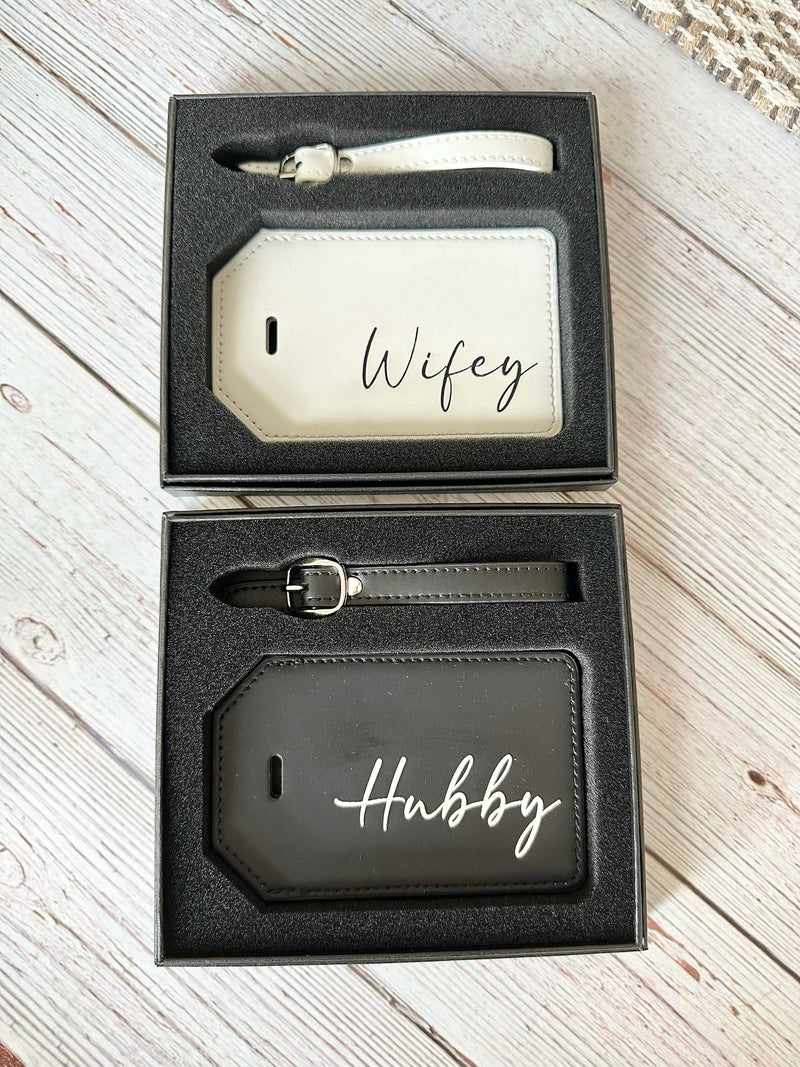 Two luggage tags with 'Wifey' and 'Hubby' engraved, in a black box on a wooden surface.